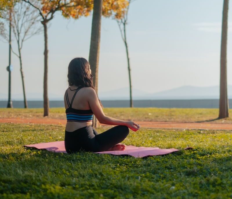 A woman meditates outdoors on a yoga mat in a tranquil park setting, surrounded by nature.