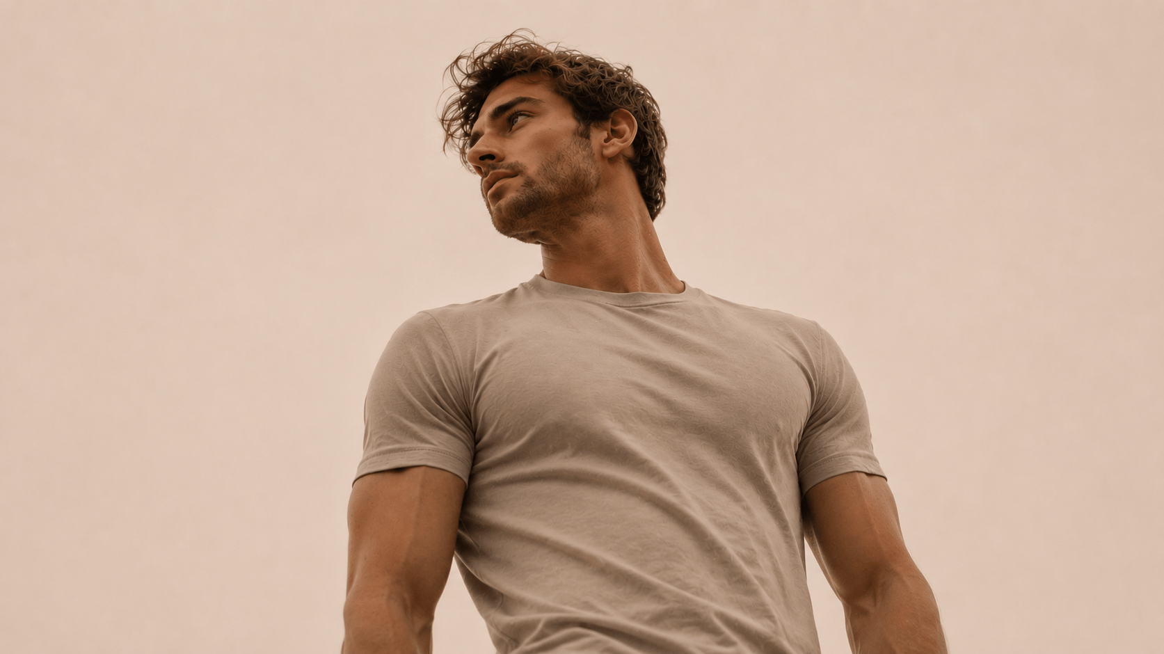 A young man with tousled hair looks confidently to the side while wearing a fitted, light-colored shirt against a soft, neutral background.